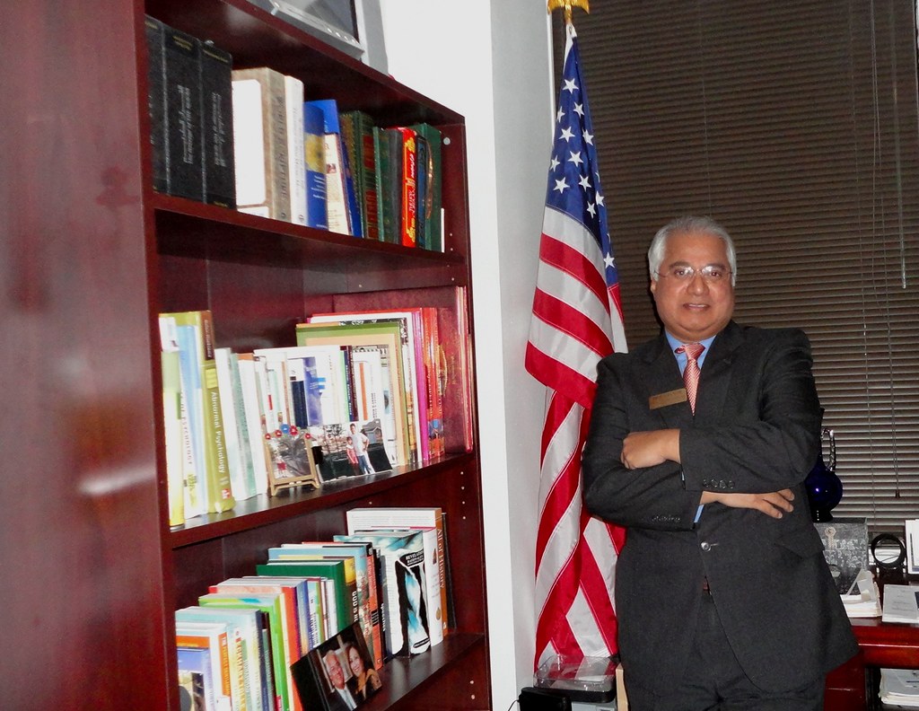 Dr. Mike Mohamed Ghouse standing beside books and an American flag, representing civic learning and pluralism